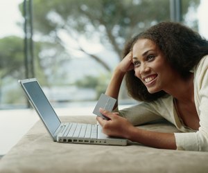 Young woman lying on bed with holding credit card and laptop, smiling, side view