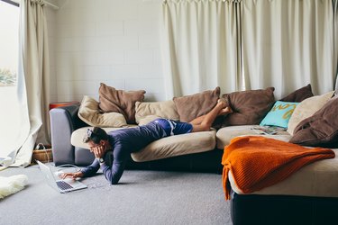 Man lying precariously on couch while reading laptop