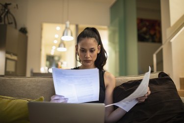 Woman reading paperwork, working at laptop on sofa