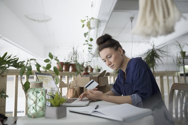 Female plant shop owner with credit card paying bills online at laptop in workshop