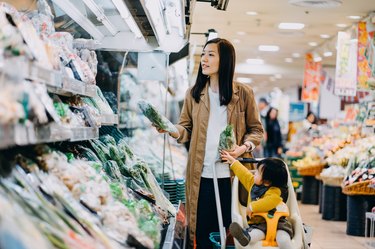 Cute little daughter sitting in a shopping cart grocery shopping for fresh organic vegetables with young Asian mother in a supermarket