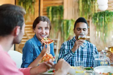 Group of joyful young friends enjoying talking, eating pizza and drinking wine during dinner in Italian restaurant