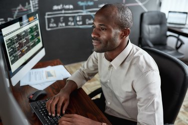 Earn more and count more. Cheerful african male trader sitting by desk and studying analytical reports using pc in the office.