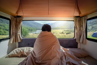 Young Asian man staying in the blanket looking at mountain scenery through the window in camper van in the morning. Road trip in summer of South Island, New Zealand.