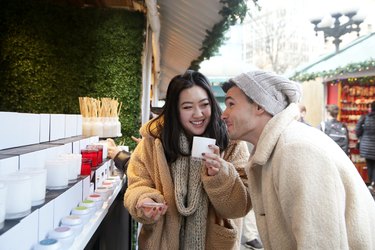 Two Millennials, one Caucasian male and one Asian female, shop for candles at outdoor holiday market in the city.