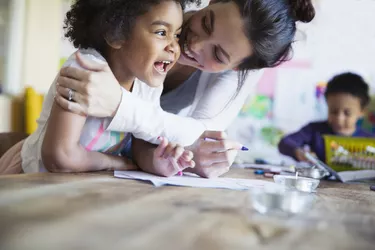 Affectionate, happy mother hugging toddler daughter coloring at table