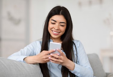 Smiling Asian Girl Browsing Social Networks On Smartphone At Home, Enjoying Weekend