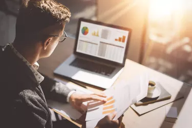 Young finance market analyst in eyeglasses working at sunny office on laptop while sitting at wooden table.Businessman analyze document in his hands.Graphs and diagramm on notebook screen.Blurred.