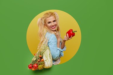 Young woman collected fresh vegetables in a string bag on a green background. Peeks out of a round hole in the wall. Retro and pin up style. The concept of proper nutrition and vegetarianism.