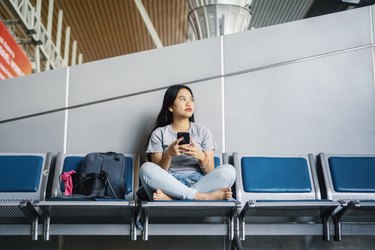 Young Asian Woman Sitting at Airport Bench Contemplating and Using Phone