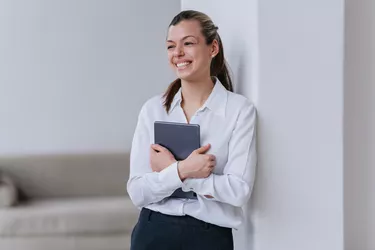 Competent young develop specialist woman standing at office holding tablet smiling wide looking aside happily. Successful manager completed all tasks. Happy student hispanic girl passed exam.