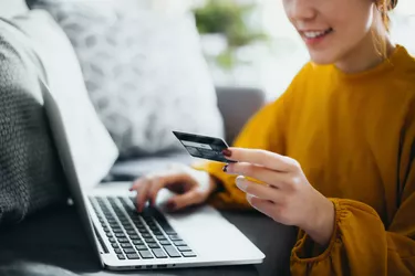 Close up of beautiful smiling young Asian woman sitting on the floor by the sofa, managing financial bill payment with laptop and credit card on hand at cozy home. Technology makes life so much easier