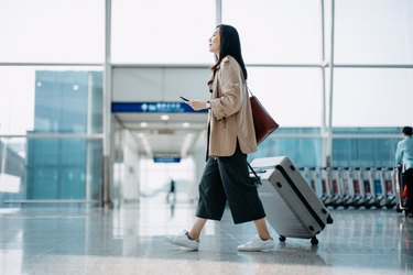Young Asian woman carrying suitcase and holding smartphone on hand, walking in airport terminal. Ready to travel. Travel and vacation concept. Business person on business trip