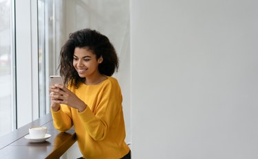 Beautiful African American woman using smartphone and mobile application for online shopping. Stylish hipster girl holding cell phone, communication, chatting, booking online, sitting in modern cafe