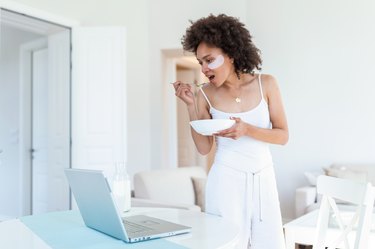 Beautiful young African American woman enjoys eating corn flakes for breakfast while working on her laptop in the morning. Looking at laptop computer and talking to her friends via video call.