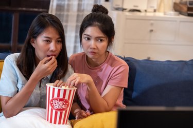 Happy asian lesbain couple watching scary movie on television together and eating popcorn at sofa in night time at home with chill ing emotion.woman friendship lifestyle