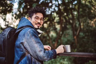 Young asian man with reusable coffee cup smiling joyfully at camera in country park