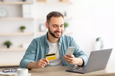 Happy Caucasian man buying things online, using smartphone, laptop and credit card, enjoying shopping in internet