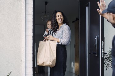 Delivery man waving to smiling female customer carrying daughter while holding paper bag at doorway