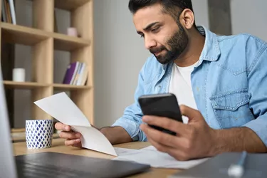 Young indian business man holding phone reading bank receipt calculating taxes.