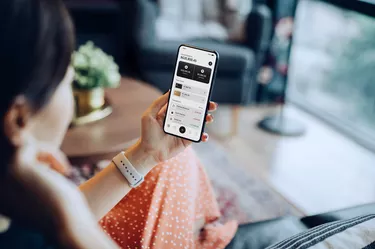 Over the shoulder view of young Asian woman relaxing at home, sitting on the sofa in the living room, managing online banking with mobile app on smartphone. Transferring money, paying bills, checking balance. Technology makes life so much easier