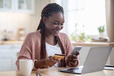 Online Payments. Cheerful black lady using smartphone and credit card in kitchen
