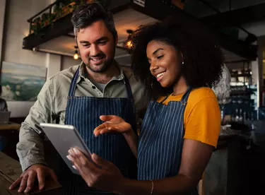 Smiling male and female colleagues discussing plans while looking at a tablet standing in coffee shop