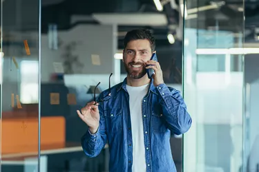 Smiling young adult bearded hipster professional business man making a business call while talking on the phone in the office, enjoying a corporate mobile conversation indoors, close up