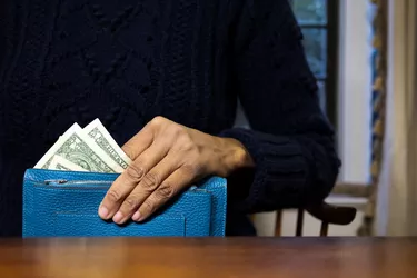 black woman holding vibrant blue wallet with multiple $1 u.s. paper bills showing while sitting at desk at home