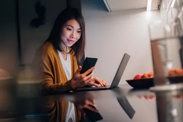 Young Woman Using Smartphone While Working With Laptop At Home In The Dark