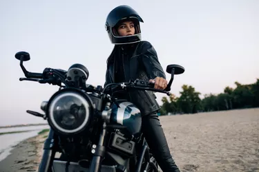 Young woman sits on motorbike in helmet on beach near river.