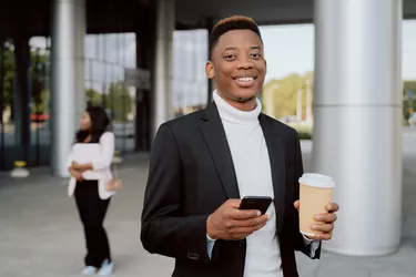 Break at work company smiling man dressed in blazer stands in front of glass-walled business center building holding phone disposable cup of take-out coffee in hand answering message