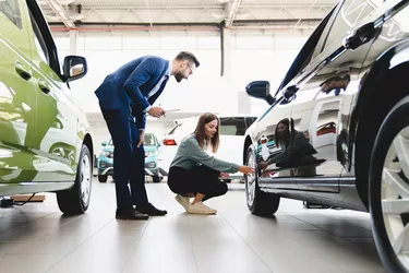 Beautiful young caucasian female client customer choosing new car, trying checking its options, tire, wheels while male shop assistant helping her to choose it at dealer auto shop