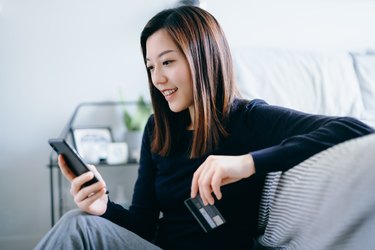 Beautiful smiling young Asian woman sitting on the floor in the bedroom, shopping online with smartphone and making mobile payment with credit card on hand at cozy home. Technology makes life so much easier