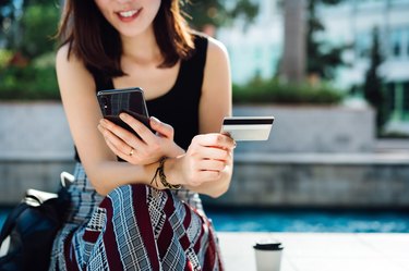 Beautiful smiling young Asian woman sitting in an urban park, enjoying coffee, shopping online with smartphone and making mobile payment with credit card on hand. Online shopping and mobile payment concept