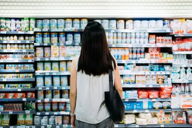 Rear view of young Asian mother groceries shopping for baby products in a supermarket. She is standing in front of the baby product aisle and have no idea which product to choose from