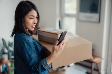 Young woman reading message on mobile phone whilst carrying a stack of delivery boxes at home