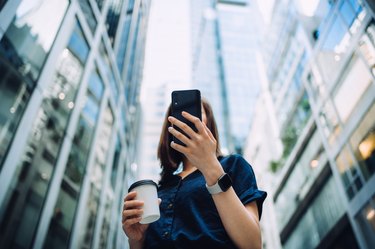 Low angle portrait of confidence and successful young Asian businesswoman with coffee to go, using smartphone while standing against highrise corporate buildings in financial district in the city