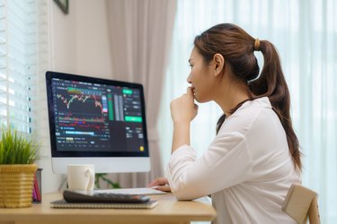 Asian woman are sitting at their desks at home trading stocks or cryptocurrency. In the living room at home. People handling various aspects of their finances from home.