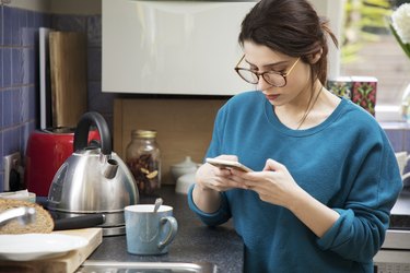 Woman looks at mobile phone while waiting for kettle to boil in kitchen.