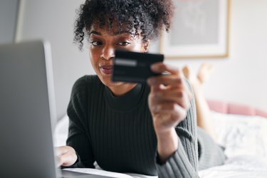 Young woman lying on bed at home with laptop computer using credit card to shop online