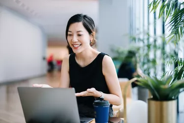 Smiling professional young Asian businesswoman talking in front of the camera having video conference with her business partners on laptop in a contemporary office space