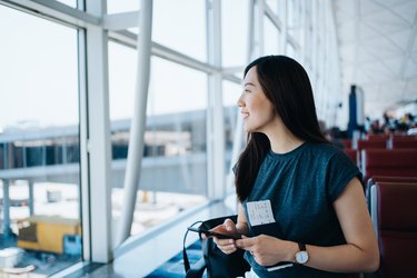 Smiling young Asian woman holding flight ticket and passport on hand, looking through the window while using smartphone and waiting for her flight in airport lounge. Travel and vacation concept. Business person on business trip