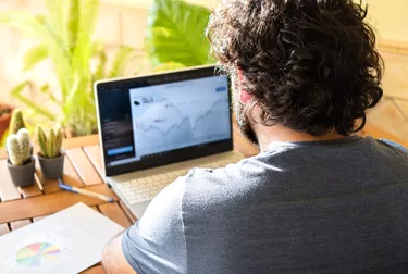 Young man sitting at table investing in stock market with laptop during work remotely from home office