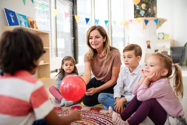 Pre- school teacher sitting on floor and playing with children indoors in nursery.