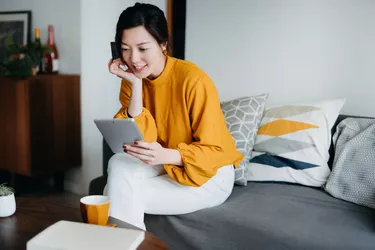 Beautiful smiling young Asian woman sitting on the sofa in the living room, shopping online with digital tablet and making mobile payment with credit card at cozy home. Technology makes life so much easier