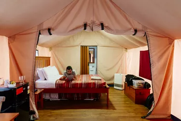 A man laying on a bed reading, inside of a 'glamping' tent