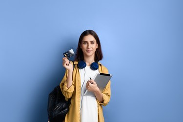 Smiling student girl is holding credit card with one hand and tablet with the other, is going to buy product and pay online, wearing yellow shirt, white t-shirt, black bag and headphones over neck.