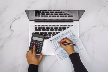 Overhead View of Hand Filling Up Tax Form and Using Calculator and Laptop