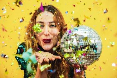 Happy fashion girl blowing confetti from hands holding a disco ball with yellow background - Young woman having fun at fest wearing trendy dress - Party, event and celebration concept - Focus on face